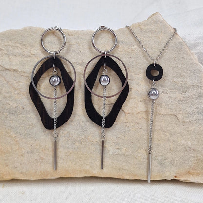 Ohrringe, Halskette und Armband aus dunkler Räuchereiche und silberfarbenen Metall liegen gemeinsam auf einem hellen Stein, die Ohrringe und die Halskette sind nebeneinander arrangiert.